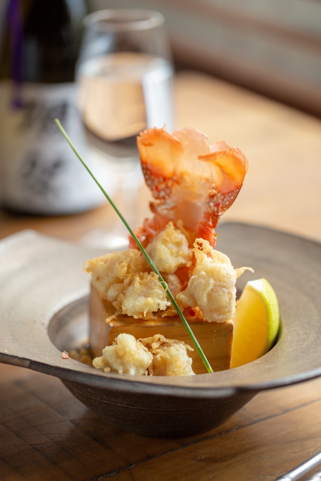 Tempura de lagosta em pedaços dourados e crocantes sobre base de madeira, com o leque da cauda erguido, fio de cebolinha e gomo de limão, em bowl rústico sobre mesa de madeira